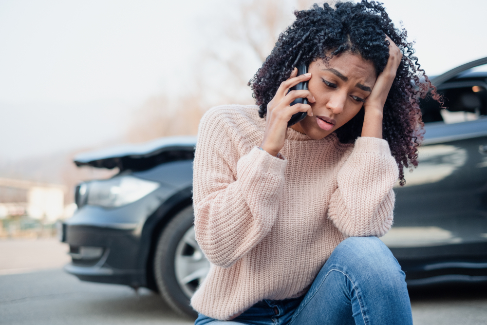 Young stressed woman on her phone with wrecked car in background.