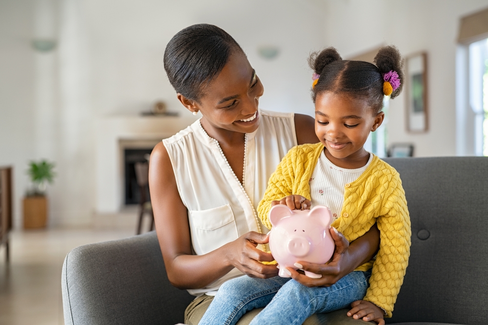 African American woman shows young child how to save money with a piggybank and bundling insurance - cheap car insurance.