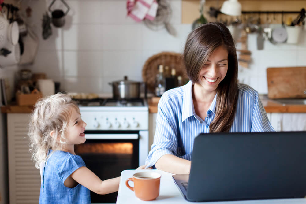 Smiling mother working freelance on her laptop with her daughter standing next to her - health insurance for freelancers.