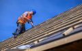 Worker installing wooden roof framing during roof replacement under clear blue sky.