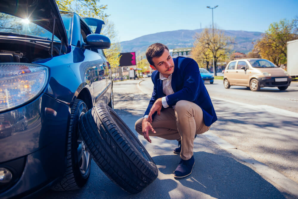 Man dressed in business clothes tries to fix a flat beside a busy road - cheap auto club.