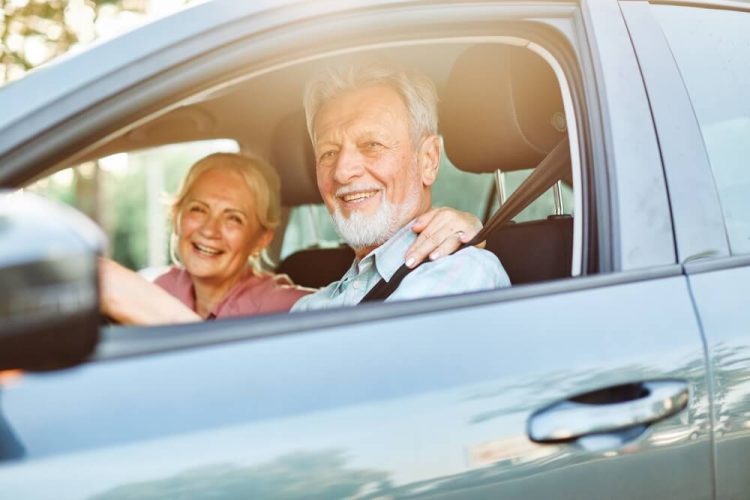 A smiling senior couple smile from inside their car, the male is driving - cheapest car insurance.