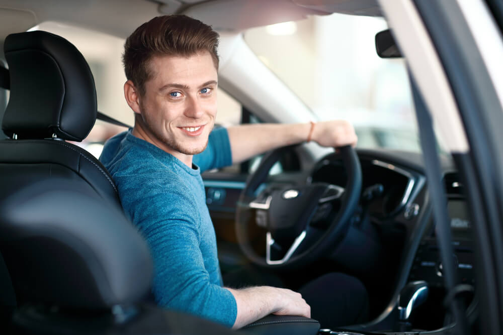 A smiling young man sits behind the wheel of his car looking back at the camera - cheapest car insurance.