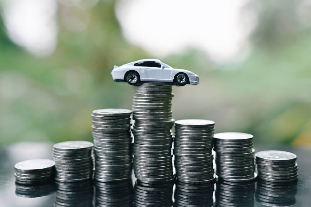 A white toy car balances on top of a stack of coins, illustrating rising car insurance rates - cheap car insurance.
