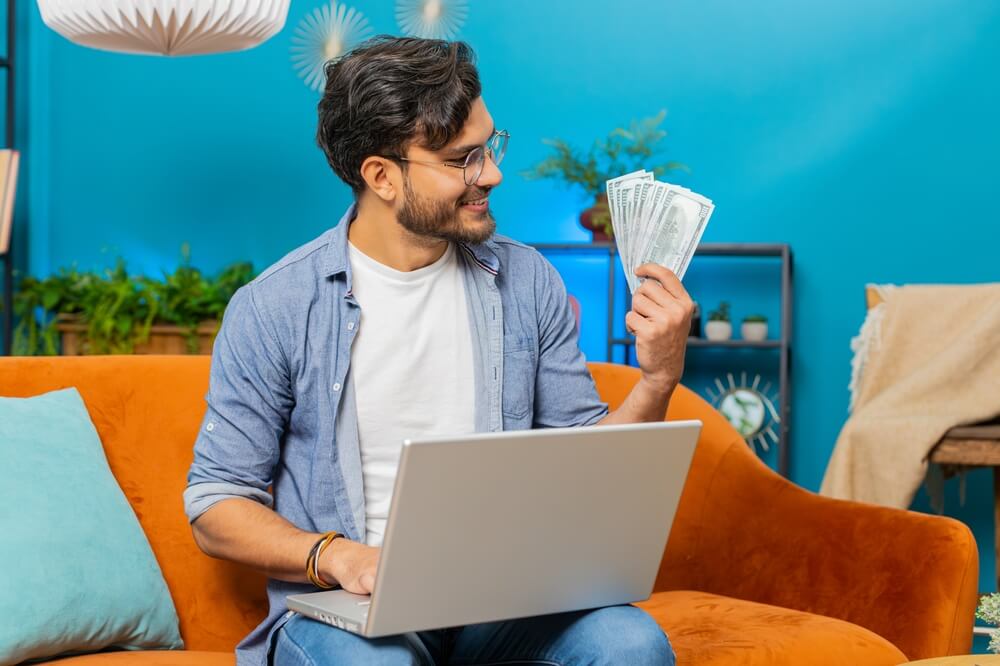 Planning budget. Rich happy Indian man counting money cash use laptop computer calculate domestic bills at home. Arabian guy satisfied of income earnings, saves money for high deductible health care.