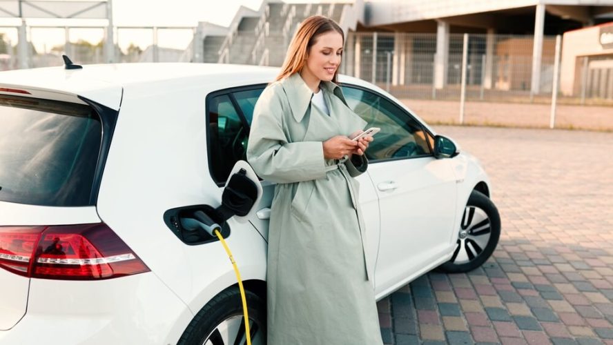 Lady stands next to her electric car while it charges at a station - cheap car insurance.