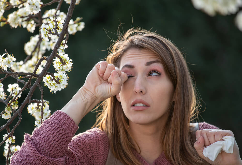 Woman rubs her eyes from spring pollen with flowers blooming - cheap car insurance.
