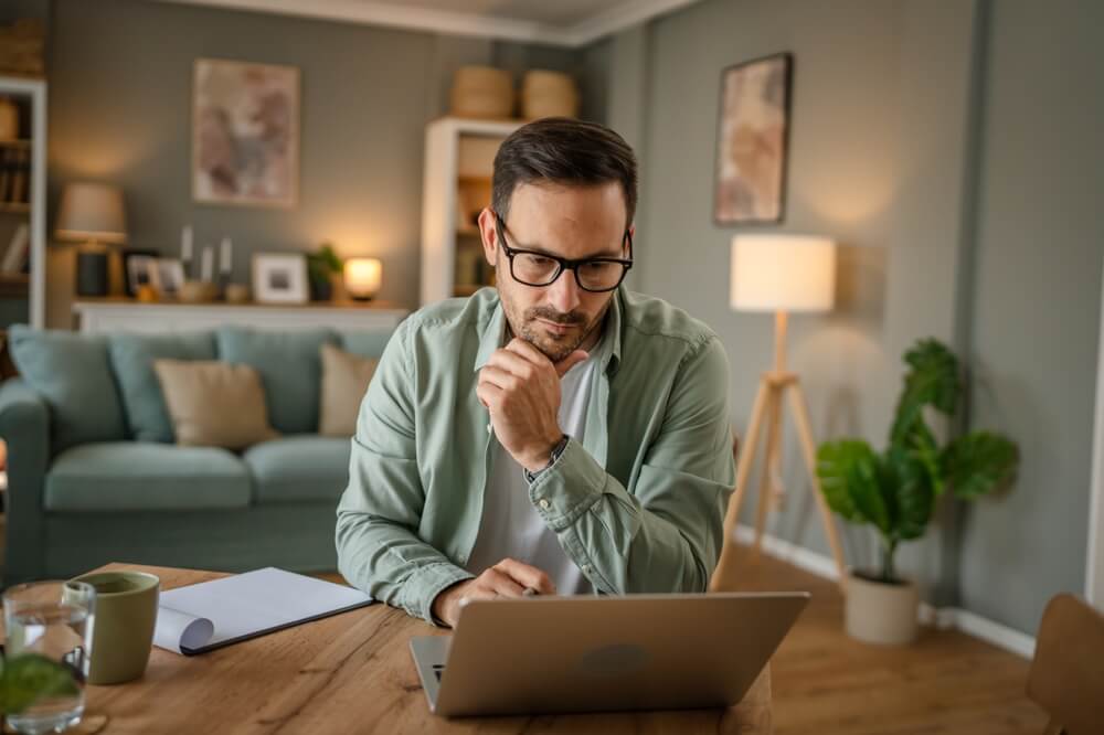 A man sits at a desk with his laptop and coffee in his living room, working from home - cheap home insurance.