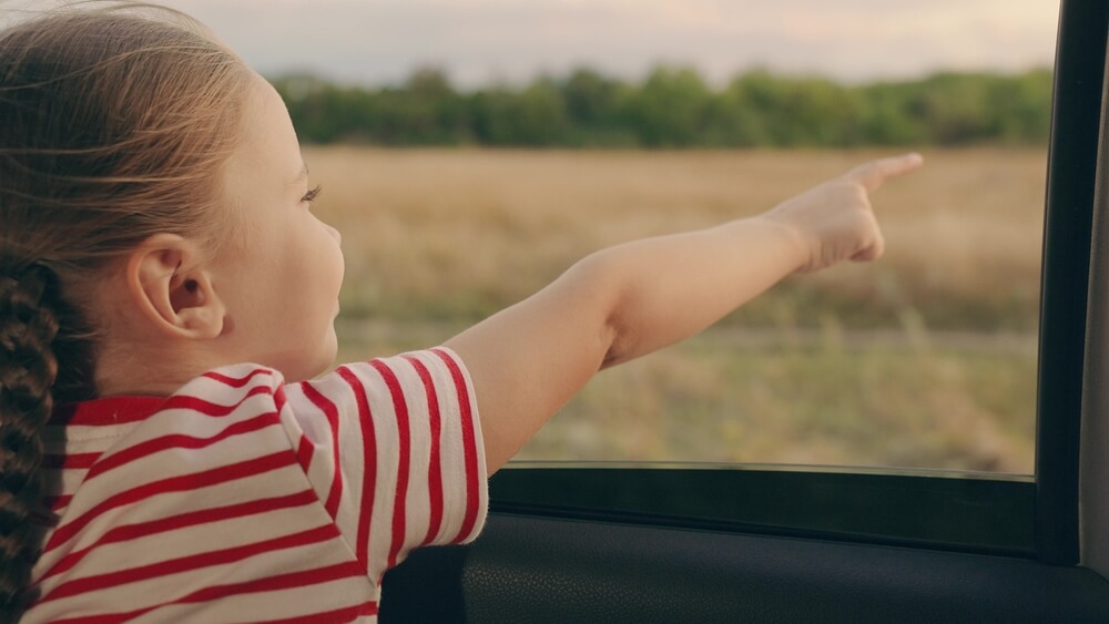 Child, stretching her hand out of car window, laughs. Girl child looks out of open car window, which keeps the car cool and uses less gas than air conditioning.