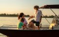 A family enjoying a boat ride with the best boat insurance near me on a calm lake during sunset. The man is standing and smiling, while the woman and child are seated and looking at him.