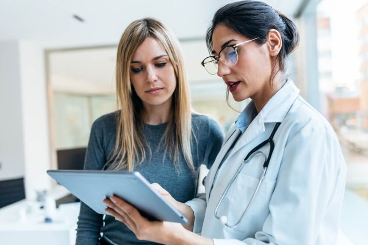 Shot of female doctor talking while explaining medical treatment with digital tablet to patient in the consultation, maximizing your Covered California health insurance.