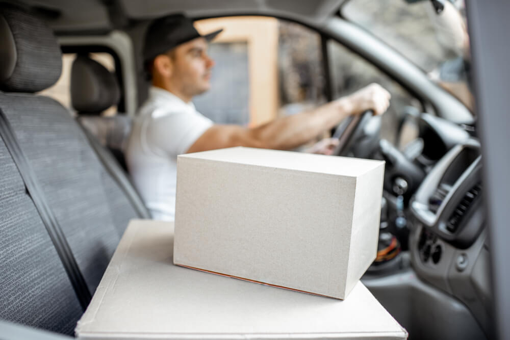 Delivery man driving cargo vehicle with parcels on the passenger seat, image focused on the cardboard boxes, importance of commercial vehicle insurance for deliveries.