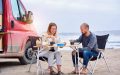 Caucasian couple sitting eating drinking coffee outdoors at the beach in van lifestyle next to their refitted van, best insurance for van lifestyle.