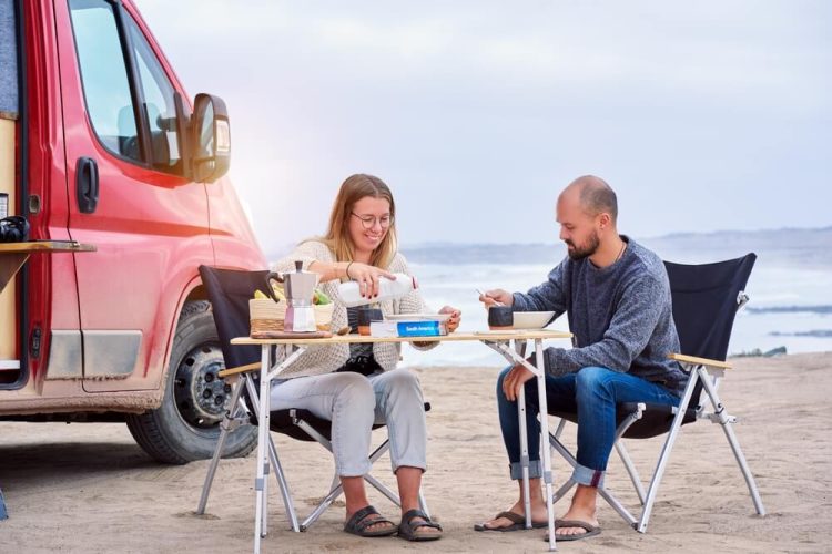 Caucasian couple sitting eating drinking coffee outdoors at the beach in van lifestyle next to their refitted van, best insurance for van lifestyle.