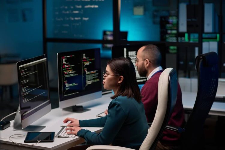 A high-angle view shows two people in a cybersecurity department, writing code under blue lights with a focus on business insurance systems.