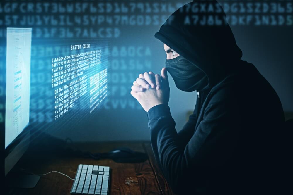 A man in a hoodie sits at his desk, coding amid blue light—a minimalist scene blending cybercrime and business insurance.