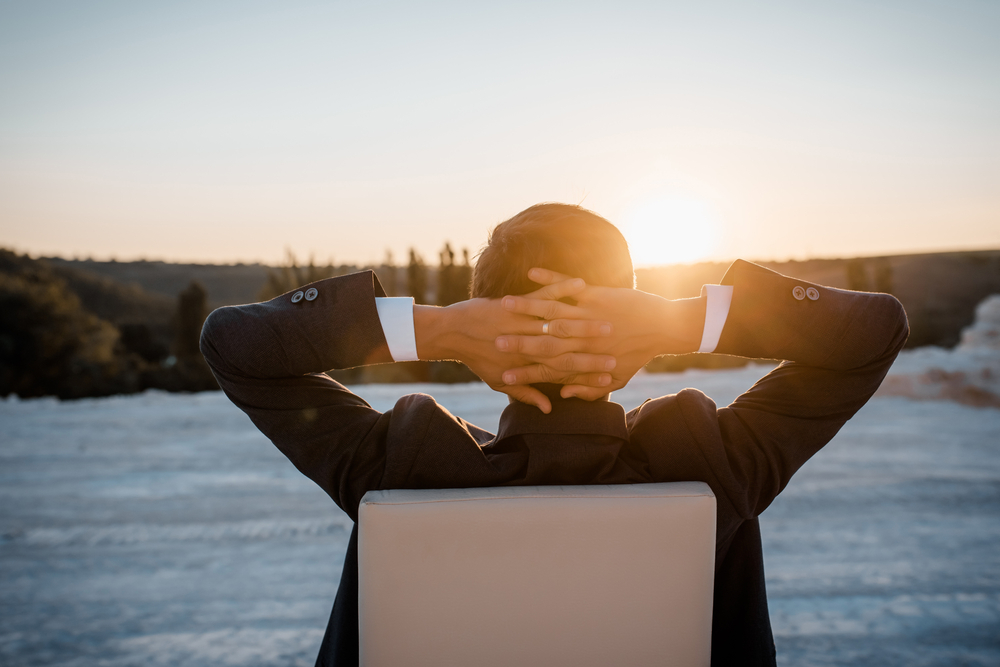 back view of young man in a classic jacket is sitting on the chair with Hands behind his head and making a rest. He is enjoying the sunset. Concept stop working, you need a vacation.