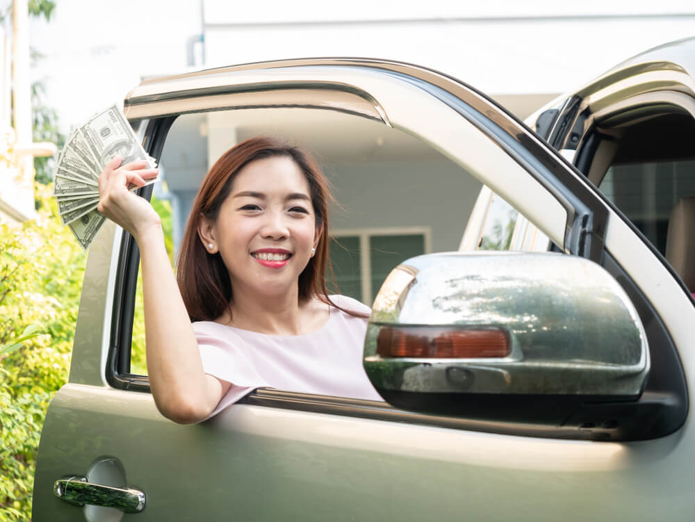 Smiling woman holding dollar bills outside her golden SUV after saving money on car insurance in big cities. 