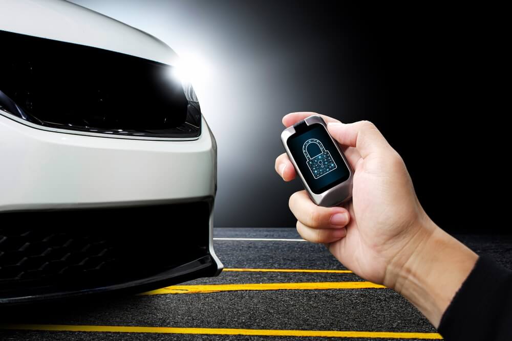 Hand holds a smart car key fob with a lock icon beside a parked vehicle, showcasing Best Car Theft Deterrent Devices like modern anti-theft alarms and immobilizers.
