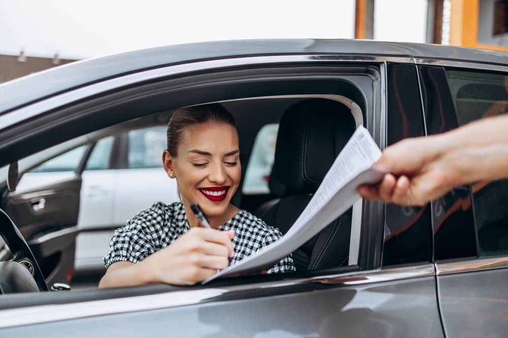 Smiling female customer sits in her newly purchased car while signing paperwork with a dealer through the open window, considering used car insurance options.