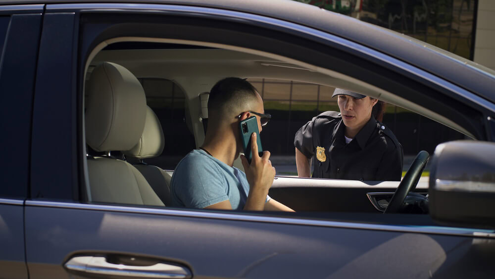 A driver holds up his smartphone while speaking with a police officer during a traffic stop, demonstrating how to provide proof of insurance on your phone.