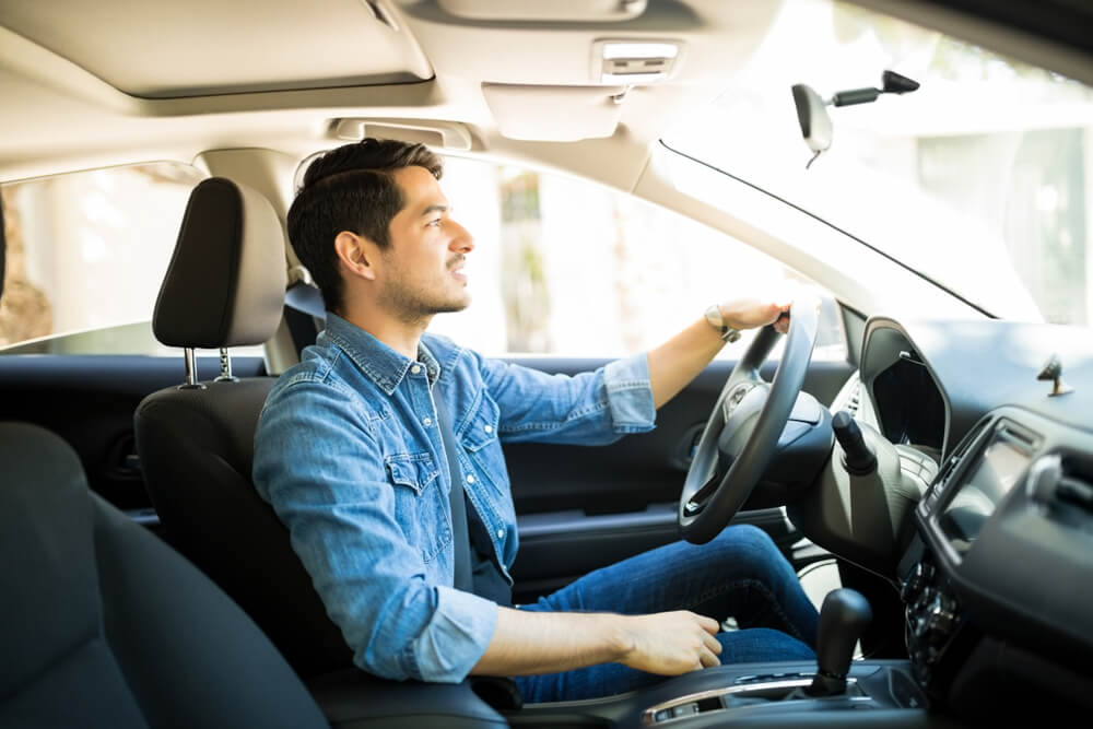 Young man in a denim shirt drives a car with a relaxed posture, representing habits that can help lower car insurance premium.