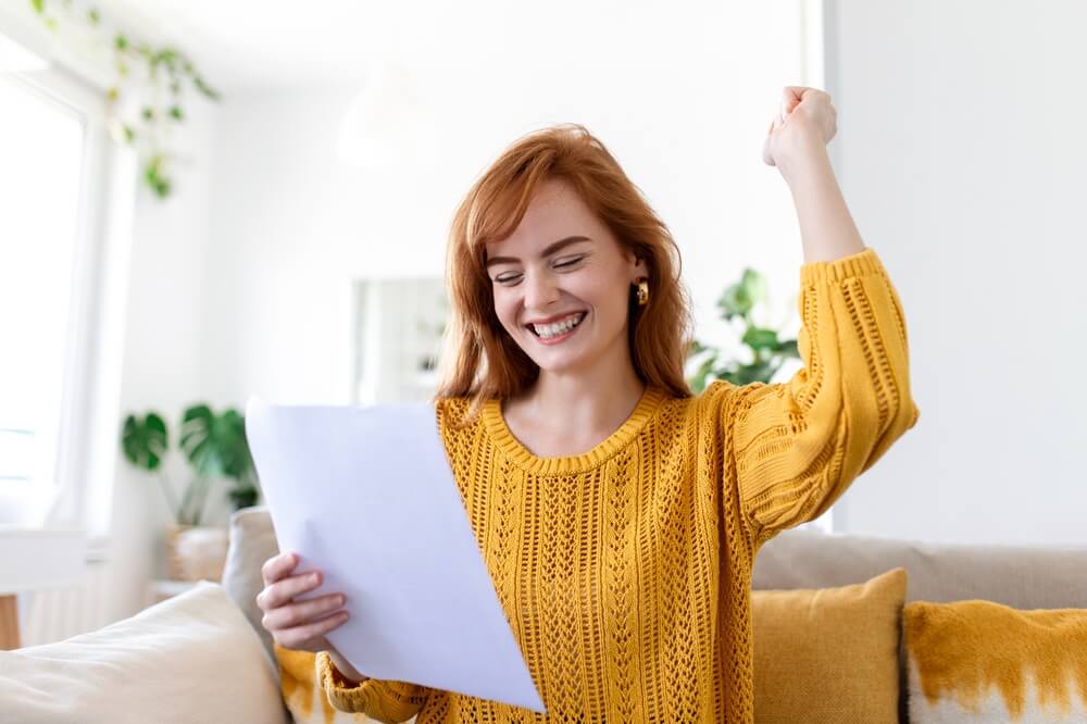 smiling red-haired woman in a yellow sweater sits on a couch holding a sheet of paper and raising her fist in celebration, suggesting savings or a refund for tax refund auto insurance.