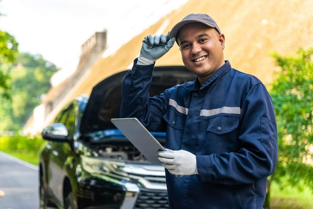Roadside mechanic in a blue uniform smiles while holding a tablet in front of a car with the hood open, explaining how to prevent car from being towed after a roadside breakdown.
