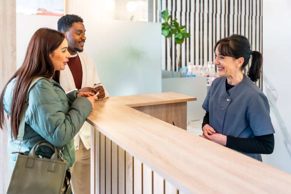 Couple speaking with a receptionist at a dental clinic, representing how dental insurance included in health insurance works.