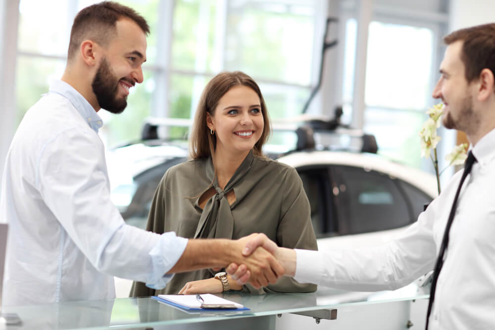 Car buyer shakes hands with a salesperson at a dealership counter while a woman smiles beside them, capturing a successful negotiation and how to beat the car salesman.