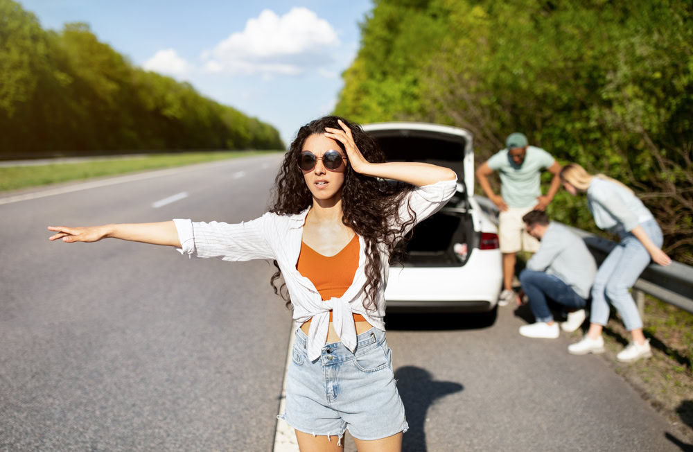 Woman signaling for roadside assistance on a highway while her car is pulled over with the trunk open and others inspect a flat tire on the shoulder.