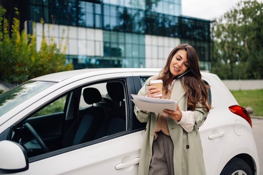 Woman standing by her car reviewing paperwork and talking on the phone while preparing to compare car insurance quotes.