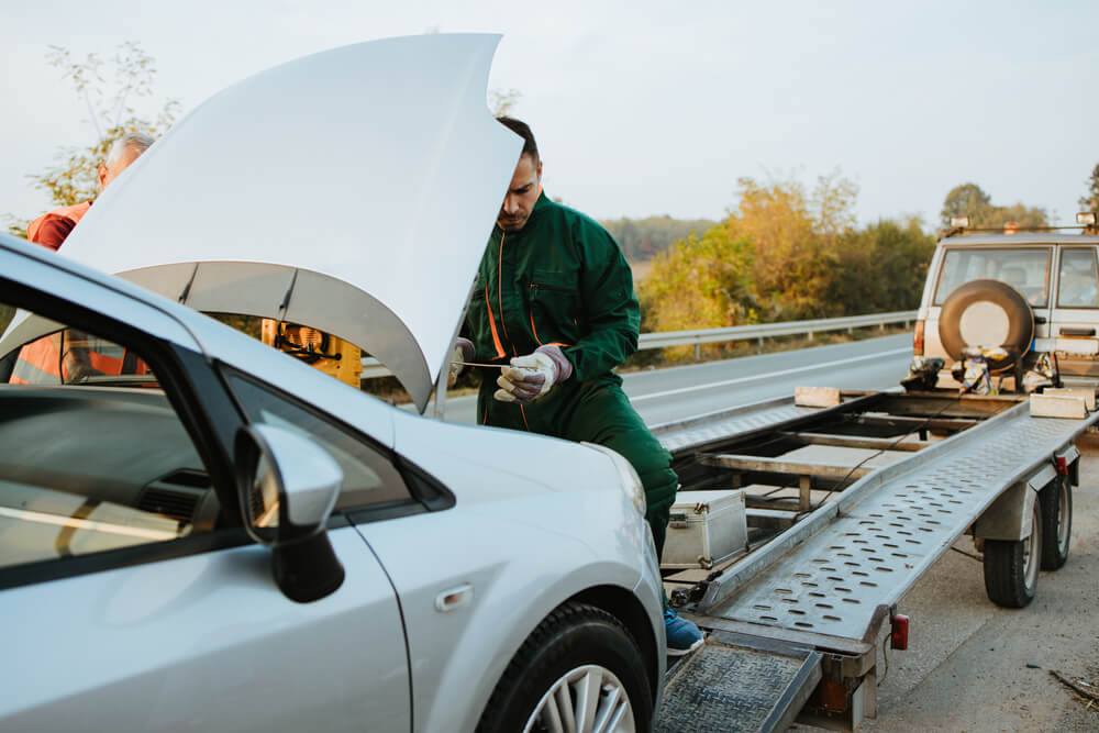 Mechanic inspecting a broken-down car with the hood open on the roadside while a tow truck prepares to load it, illustrating the importance of insurance add-ons like roadside assistance, towing coverage, and emergency breakdown protection.