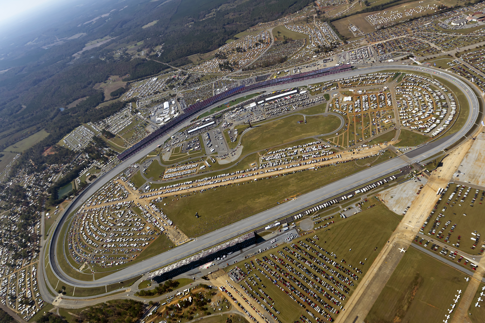 Aerial view of Talladega Superspeedway showing the large oval NASCAR track surrounded by packed grandstands, parking lots filled with cars and RVs, and the infield area bustling with activity.