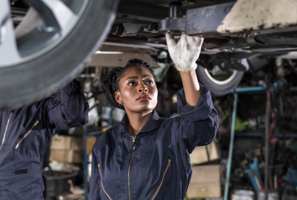 Black auto mechanic woman in overall and working gloves fixing car in auto repair garage.
