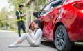 Woman sitting beside a red car with major side damage after an accident while an insurance adjuster documents the scene, illustrating a car insurance record claim.