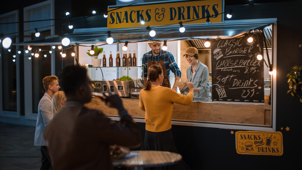 Customers order food and drinks from a brightly lit food truck at night while staff members serve guests through the service window under string lights