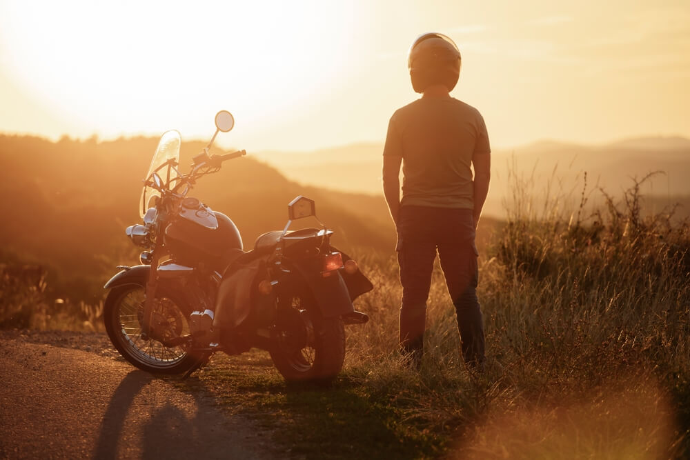 Motorcyclist wearing a helmet stands beside a parked bike at sunset, highlighting Motorcycle Helmets and Insurance.
