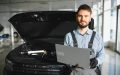 Certified technician using a laptop to diagnose vehicle software repairs with the hood open in a professional service center.