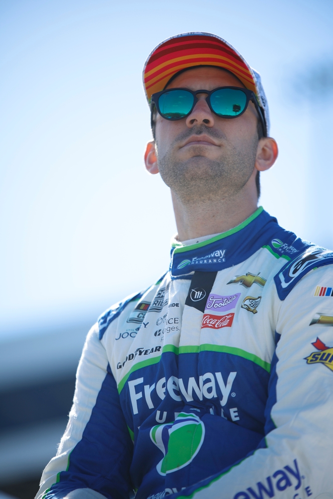 NASCAR driver wearing sunglasses, a racing suit, and a colorful cap poses outdoors under a bright blue sky.