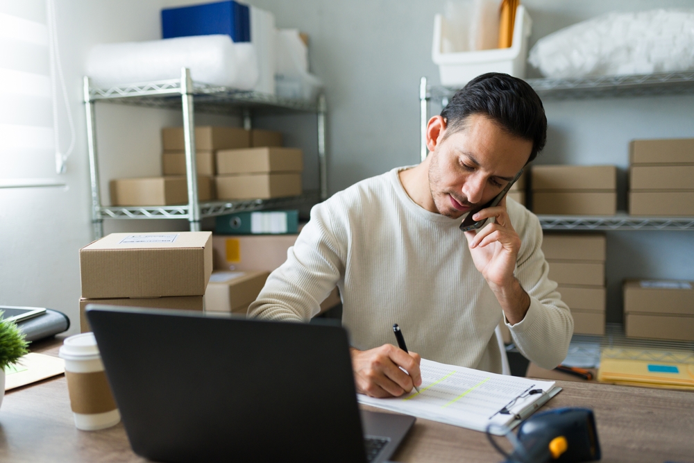 Small business owner talking on the phone while reviewing paperwork at a desk surrounded by shipping boxes.