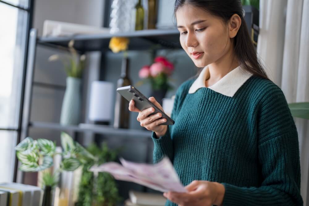 Woman reviewing bills and checking her phone while deciding to pay in full or monthly for insurance.