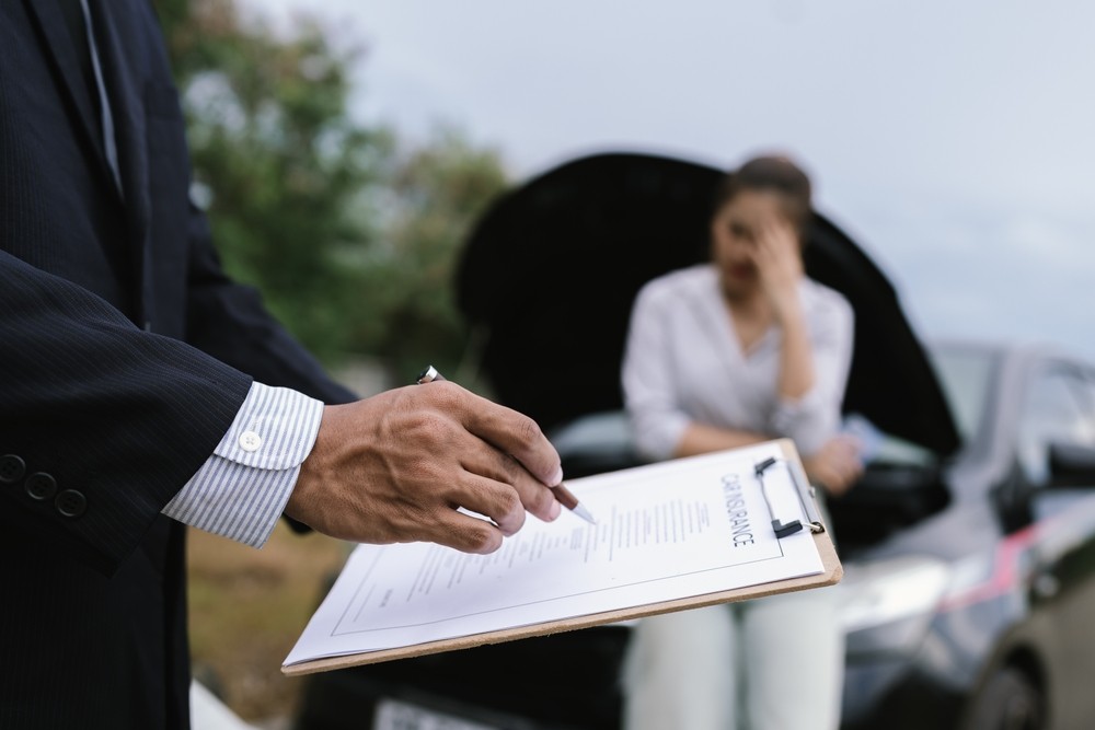 Insurance representative reviewing an auto insurance claim while a worried driver stands beside a broken-down car.