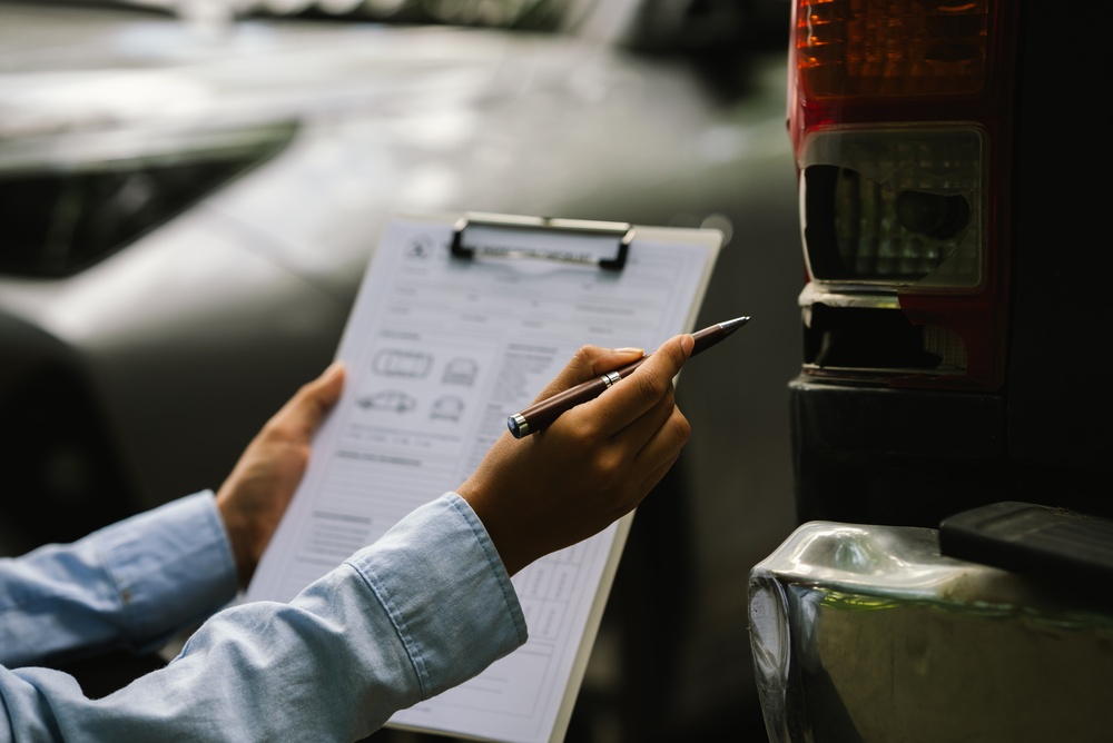 Person holding a clipboard and pen while completing a car insurance claim form beside a vehicle.