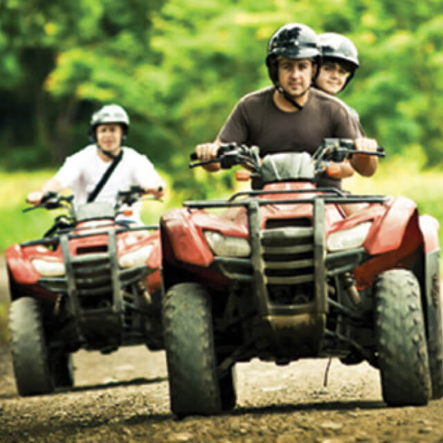 Three people riding two ATVs on a dirt road.