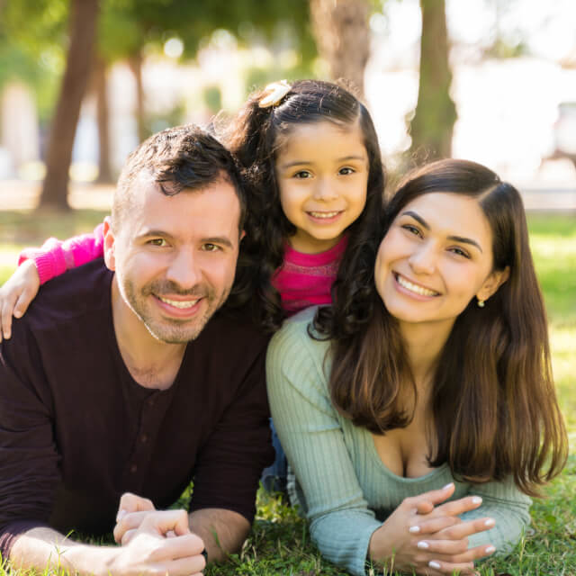 A smiling family composed by father, mother and a young girl lying on grass.