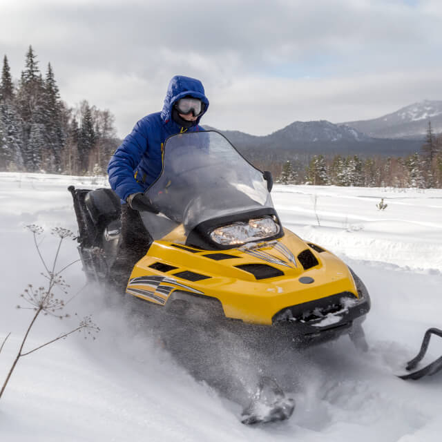 A man riding a snowmobile with a forest and mountains in the background.