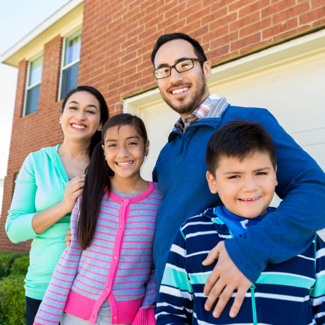 A family composed by a man, a woman, a boy and a girl smiling in front of a house.