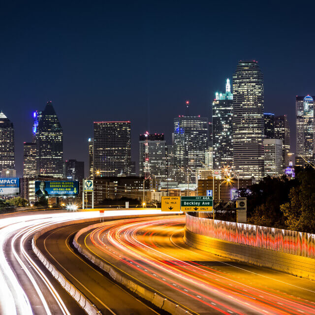 Picture of Dallas city at night including a main road with high buildings in background.