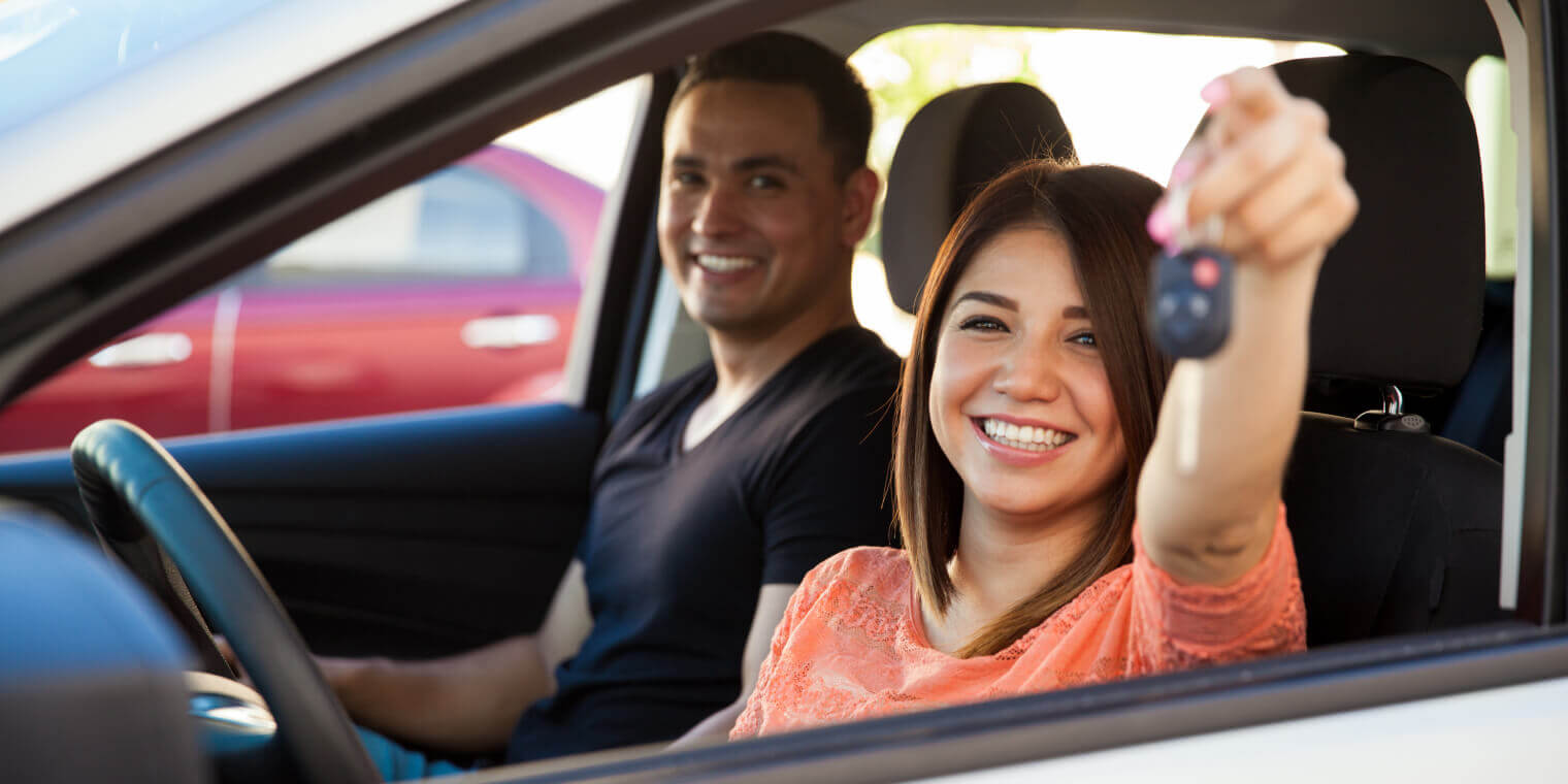 Smiling woman in a car driver's seat holding keys up with a man as passenger